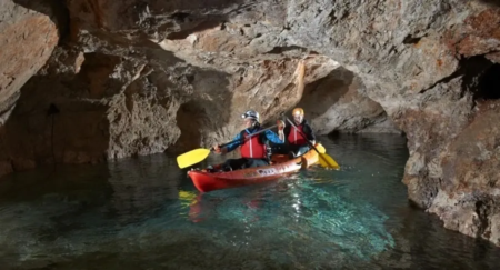 Two people kayaking through an underground cave with clear water and rocky walls, wearing helmets and life jackets.