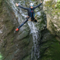 A person wearing a wetsuit, helmet, and harness jumps mid-air beside a small waterfall, surrounded by mossy rock formations