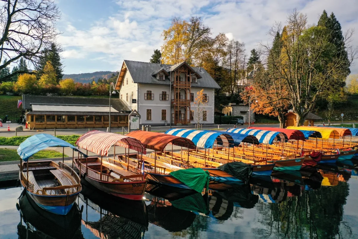 Colorful boats with striped canopies moored by a calm lake, with an Alpine-style building and autumn trees in the background.