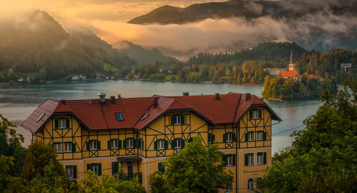 Charming yellow building overlooking Lake Bled, with a misty sunrise and an island church in the background.