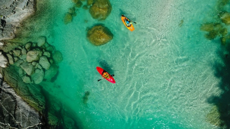 Aerial view of kayakers paddling on the clear turquoise Soča River with rocks visible underwater.