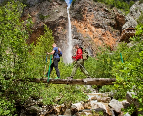 Hikers crossing rocks near Rinka Waterfall in Logar Valley surrounded by forest and cliffs