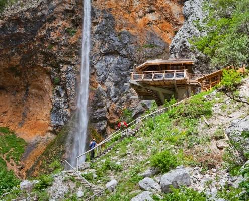 Hikers crossing rocks near Rinka Waterfall in Logar Valley surrounded by forest and cliffs